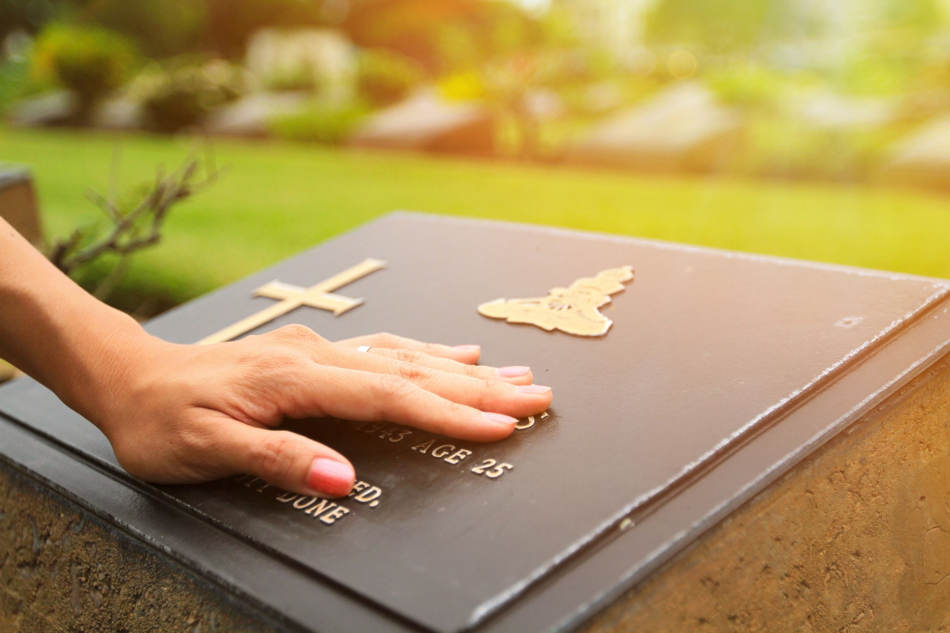 Woman's hand touching the black stone grave at cemetery with orange sunlight flare.